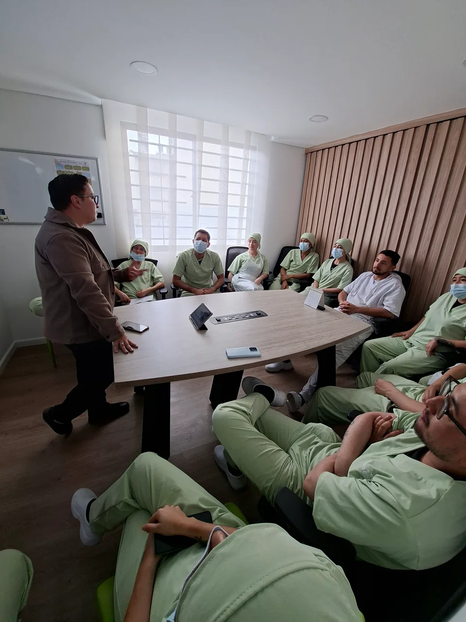 Instructor explica un tema de seguridad y salud en el trabajo a personal de salud sentado alrededor de una mesa.