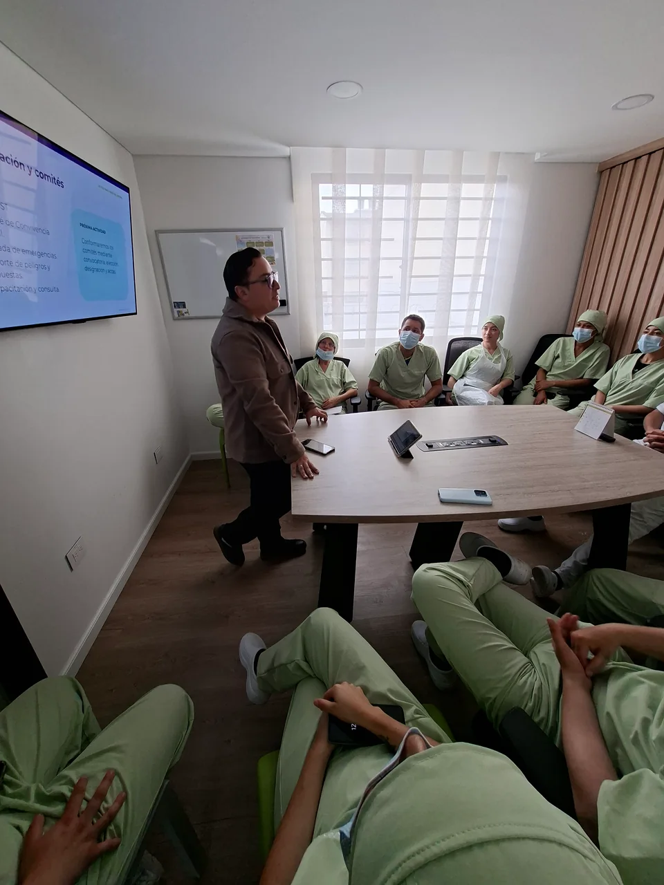Instructor dirige una capacitación presencial con una pantalla de apoyo y personal de salud alrededor de una mesa.